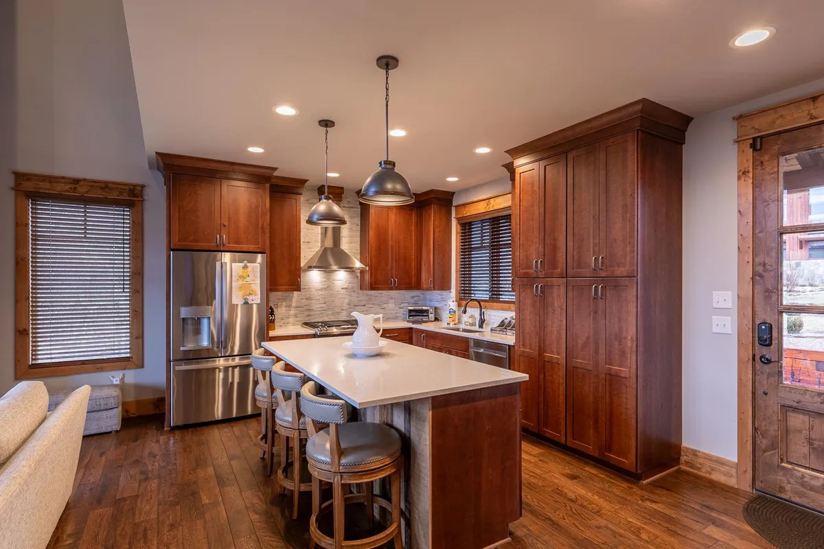 This image presents the heart of the kitchen, showcasing a central island with seating, stainless steel appliances, warm wood cabinetry, and stylish pendant lighting. The inviting setup suggests ample space for collaborative cooking, casual meals, and socializing.