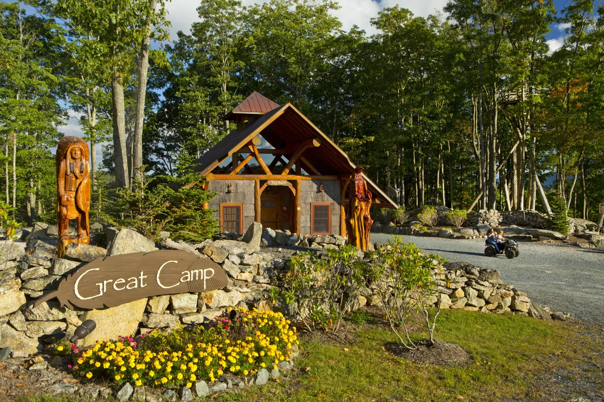 The image highlights the 'Great Camp' sign, whimsical wooden sculptures, main cabin structure, and flower beds. The scene feels lively and welcoming, suggesting a hub for family activities, games, and exploration, surrounded by natural beauty.