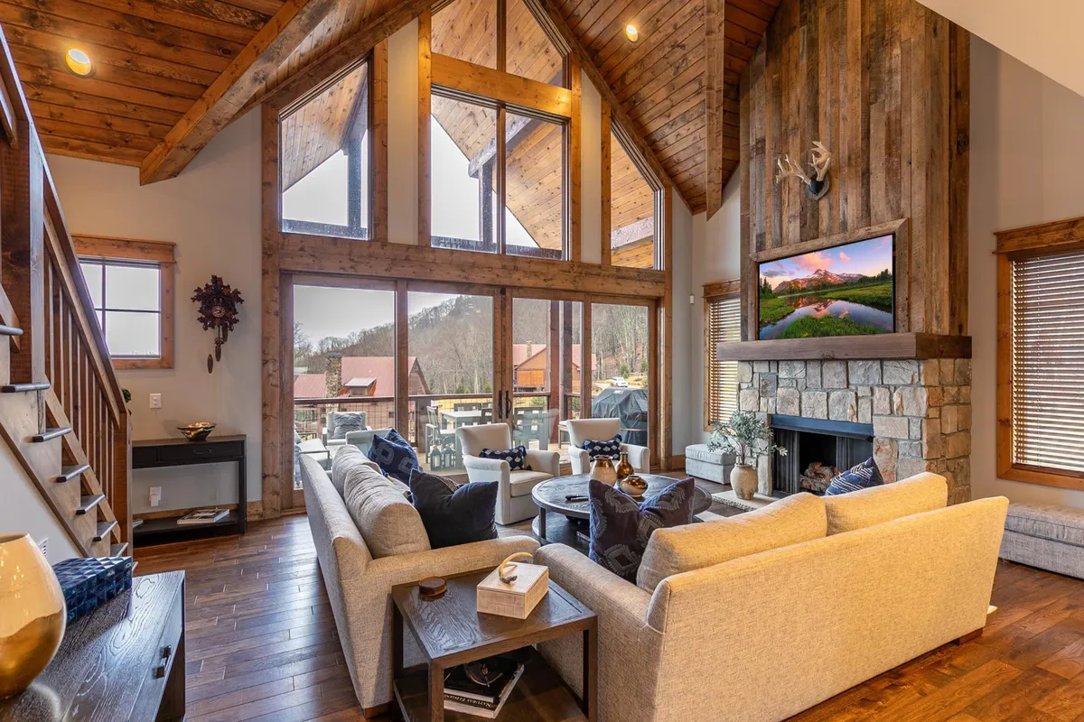 A spacious great room filled with natural light from large windows, featuring a dramatic stone fireplace as its centerpiece, exposed wooden beams, comfortable seating, and a TV mounted above the fireplace. The room exudes warmth and invites gatherings, with its open-plan layout seamlessly connecting the living area to adjacent spaces.