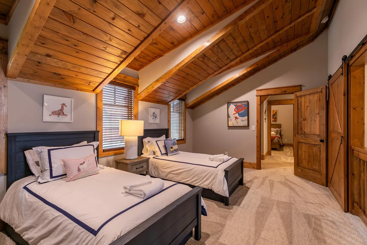 A loft-style room with two twin beds, warm wooden beams, a table lamp, and playful artwork. The space feels welcoming and flexible—perfect for kids or friends to enjoy sleepovers or private moments.