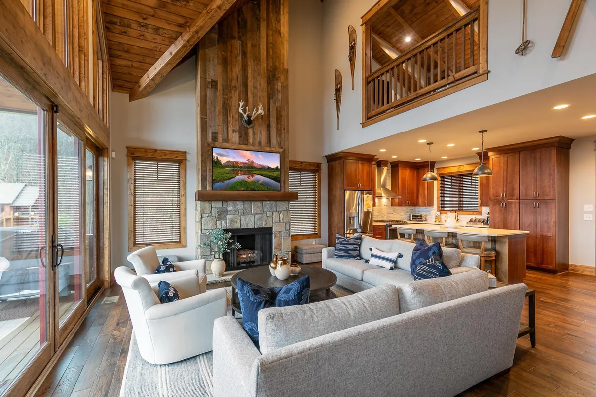 A wide-angle view of the open-plan layout, featuring the stone fireplace, large windows, soaring wooden beams, and a glimpse into the connected kitchen area. The space feels expansive yet inviting, perfectly designed for groups to share meals, relax, and socialize without barriers.