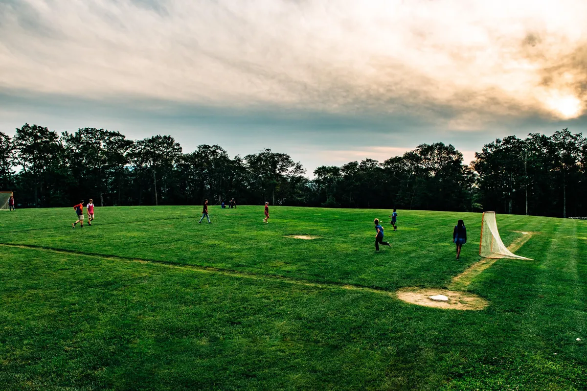 Children are seen playing soccer and lacrosse on a lush green field surrounded by trees, illustrating the playful, energetic side of the community's outdoor amenities and the family-friendly environment.